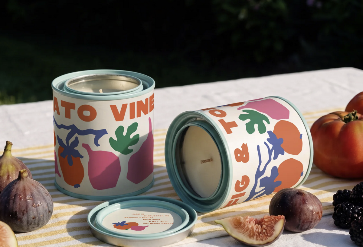 Two decorative metal candle tins with a colorful botanical and fruit print, styled on a striped tablecloth with fresh figs and tomatoes in an outdoor setting.